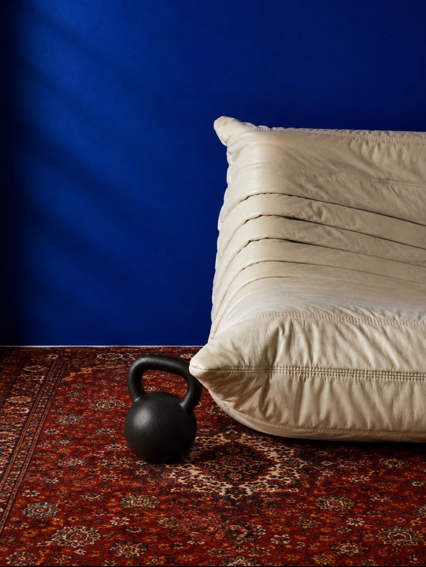 Cream leather bench and a kettlebell on a red patterned rug against a blue backdrop